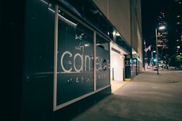A city street at night with a modern glass building facade showcasing a large management company logo. The area is illuminated by streetlights, and a parking garage entrance is visible on the right. The street is quiet, with some lights visible in the windows of a large building in the background.