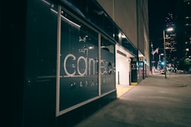 A city street at night with a modern glass building facade showcasing a large management company logo. The area is illuminated by streetlights, and a parking garage entrance is visible on the right. The street is quiet, with some lights visible in the windows of a large building in the background.