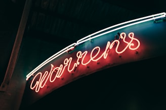 A glowing neon sign displaying cursive letters in an orange hue, set against a dark background. The neon tubes are bright and create a warm ambiance, with a hint of blue and white light accentuating the top.