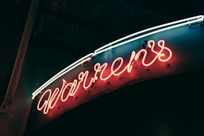 A glowing neon sign displaying cursive letters in an orange hue, set against a dark background. The neon tubes are bright and create a warm ambiance, with a hint of blue and white light accentuating the top.