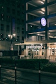 An urban street scene at night featuring a building with a parking garage. A bright illuminated sign of a restaurant named 'sweetgreen' is visible on the ground floor. The area is lit by streetlights, and the atmosphere is calm and quiet.