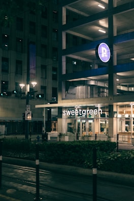 An urban street scene at night featuring a building with a parking garage. A bright illuminated sign of a restaurant named 'sweetgreen' is visible on the ground floor. The area is lit by streetlights, and the atmosphere is calm and quiet.