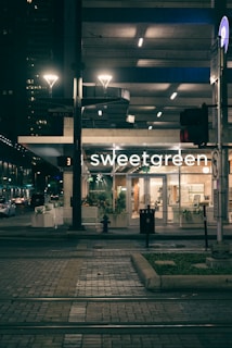 An evening urban scene featuring a well-lit storefront with the name 'sweetgreen'. The surroundings include a tram track in the foreground, illuminated streetlights, and an urban background with other buildings visible in dim lighting. Potted plants are placed in front of the store, contributing to a modern and inviting atmosphere.