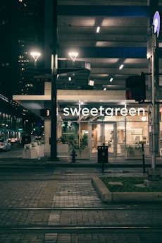 An evening urban scene featuring a well-lit storefront with the name 'sweetgreen'. The surroundings include a tram track in the foreground, illuminated streetlights, and an urban background with other buildings visible in dim lighting. Potted plants are placed in front of the store, contributing to a modern and inviting atmosphere.