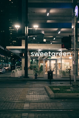 An evening urban scene featuring a well-lit storefront with the name 'sweetgreen'. The surroundings include a tram track in the foreground, illuminated streetlights, and an urban background with other buildings visible in dim lighting. Potted plants are placed in front of the store, contributing to a modern and inviting atmosphere.