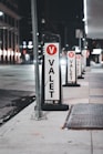 Several valet signs are lined up along a city street at night. The signs are marked with a large red V and the word 'VALET' in bold black letters on a white background. The street is dimly lit, with buildings and streetlights visible in the background.
