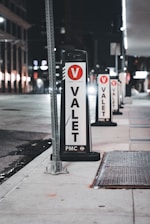 Several valet signs are lined up along a city street at night. The signs are marked with a large red V and the word 'VALET' in bold black letters on a white background. The street is dimly lit, with buildings and streetlights visible in the background.