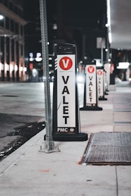 Several valet signs are lined up along a city street at night. The signs are marked with a large red V and the word 'VALET' in bold black letters on a white background. The street is dimly lit, with buildings and streetlights visible in the background.