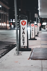 Several valet signs are lined up along a city street at night. The signs are marked with a large red V and the word 'VALET' in bold black letters on a white background. The street is dimly lit, with buildings and streetlights visible in the background.