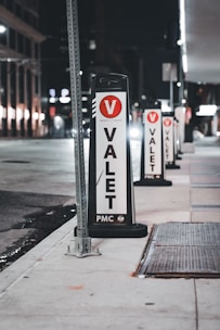 Several valet signs are lined up along a city street at night. The signs are marked with a large red V and the word 'VALET' in bold black letters on a white background. The street is dimly lit, with buildings and streetlights visible in the background.
