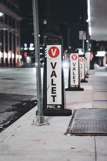 Several valet signs are lined up along a city street at night. The signs are marked with a large red V and the word 'VALET' in bold black letters on a white background. The street is dimly lit, with buildings and streetlights visible in the background.