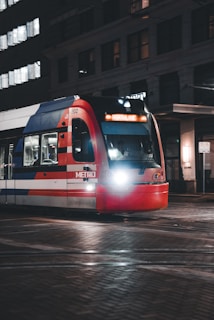 A city light rail train, identified by the 'Metro' label on its side, travels along tracks in an urban area during nighttime. The train displays 'Northline' on its digital destination board and is illuminated by its headlights. Surrounding buildings are partly visible with some windows lit.