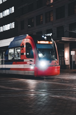 A city light rail train, identified by the 'Metro' label on its side, travels along tracks in an urban area during nighttime. The train displays 'Northline' on its digital destination board and is illuminated by its headlights. Surrounding buildings are partly visible with some windows lit.