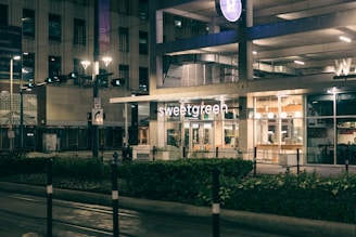 A modern urban environment featuring the storefront of a Sweetgreen restaurant. The building exterior is illuminated at night with glass windows revealing interior lighting and seating. Surrounding the building are streetlights, traffic signs, and a well-maintained sidewalk with greenery.