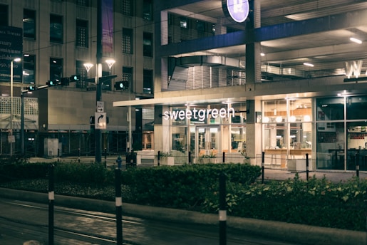 A modern urban environment featuring the storefront of a Sweetgreen restaurant. The building exterior is illuminated at night with glass windows revealing interior lighting and seating. Surrounding the building are streetlights, traffic signs, and a well-maintained sidewalk with greenery.