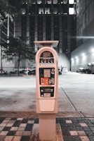 A parking meter stands in an urban setting at night, surrounded by buildings and a faintly lit parking area. The tall structure behind the meter has multiple windows illuminated, indicating occupancy. The street is lined with patterned tiles leading to the meter, and a few trees are present in the background.