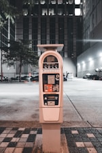 A parking meter stands in an urban setting at night, surrounded by buildings and a faintly lit parking area. The tall structure behind the meter has multiple windows illuminated, indicating occupancy. The street is lined with patterned tiles leading to the meter, and a few trees are present in the background.