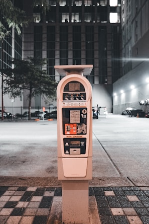 A parking meter stands in an urban setting at night, surrounded by buildings and a faintly lit parking area. The tall structure behind the meter has multiple windows illuminated, indicating occupancy. The street is lined with patterned tiles leading to the meter, and a few trees are present in the background.