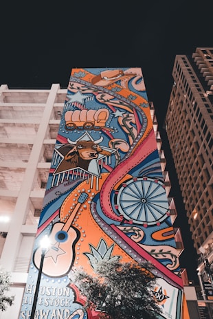 A vibrant mural on a tall building features colorful illustrations of western motifs including a wagon, a longhorn steer, a guitar, stars, and a Ferris wheel. The artwork is filled with swirling patterns and bold lines, bringing a lively and eclectic aesthetic against the backdrop of an urban setting.