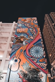 A vibrant mural on a tall building features colorful illustrations of western motifs including a wagon, a longhorn steer, a guitar, stars, and a Ferris wheel. The artwork is filled with swirling patterns and bold lines, bringing a lively and eclectic aesthetic against the backdrop of an urban setting.
