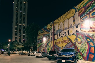 A vibrant street mural reads 'Houston is Inspired' with colorful patterns and shapes surrounding the text. Parked cars, including a blue Chevrolet, are visible in front of the mural. A tall building and street lamps illuminate the scene, creating a lively urban atmosphere at night.