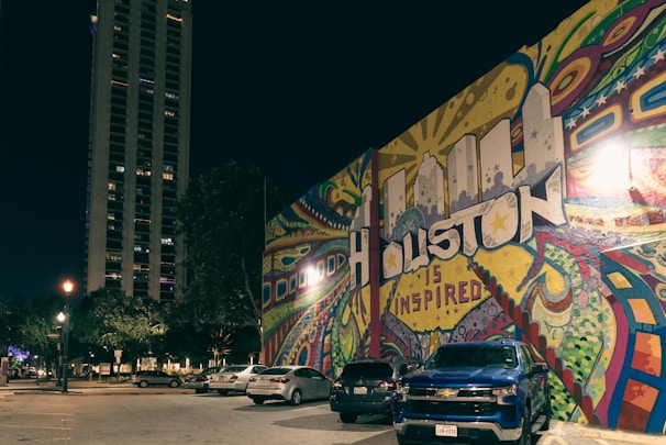A vibrant street mural reads 'Houston is Inspired' with colorful patterns and shapes surrounding the text. Parked cars, including a blue Chevrolet, are visible in front of the mural. A tall building and street lamps illuminate the scene, creating a lively urban atmosphere at night.