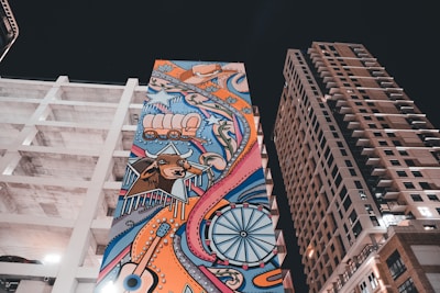A tall, colorful mural is painted on the side of a building, featuring a variety of imagery including a bull's head, a covered wagon, and a star. The artwork incorporates swirling patterns and vibrant colors, set against a night sky. The surrounding area includes modern, mid-rise buildings with geometric architectural designs.