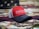 A red and black baseball cap with the words 'SAWGRASS FISHING RODS' embroidered on the front. The background features a blurred American flag theme, and there are fishing lures partially visible around the hat.