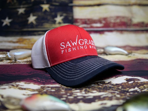 A red and black baseball cap with the words 'SAWGRASS FISHING RODS' embroidered on the front. The background features a blurred American flag theme, and there are fishing lures partially visible around the hat.
