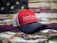A red and black baseball cap with the words 'SAWGRASS FISHING RODS' embroidered on the front. The background features a blurred American flag theme, and there are fishing lures partially visible around the hat.