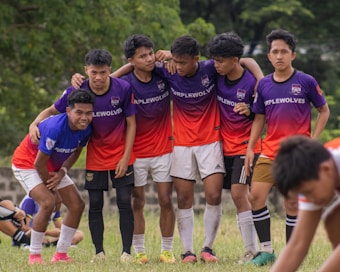 A group of young men standing closely together on a grassy field, wearing matching soccer jerseys that say 'Purplewolves'. They appear to be in a team huddle, displaying camaraderie and teamwork. A few of them are smiling, with arms around each other. The setting is outdoors with green trees in the background.
