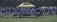 A group of football players in uniform standing in a line on a football field. They are wearing light blue jerseys, and some have helmets in their hands. A tent and goal nets are visible in the background.