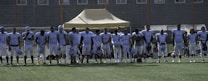A group of football players in uniform standing in a line on a football field. They are wearing light blue jerseys, and some have helmets in their hands. A tent and goal nets are visible in the background.