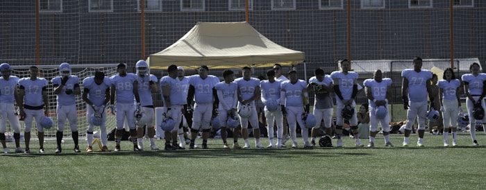 A group of football players in uniform standing in a line on a football field. They are wearing light blue jerseys, and some have helmets in their hands. A tent and goal nets are visible in the background.
