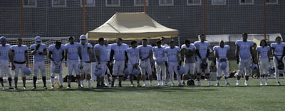 A group of football players in uniform standing in a line on a football field. They are wearing light blue jerseys, and some have helmets in their hands. A tent and goal nets are visible in the background.