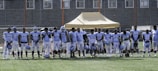 A group of football players wearing matching white and light blue uniforms stand in a line on a grassy field. They are holding helmets in their hands, and appear to be next to a beige tent structure. The background shows a wire fence and buildings.