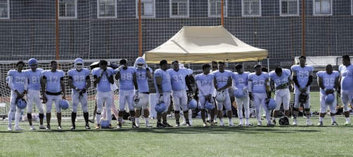 A group of football players wearing matching white and light blue uniforms stand in a line on a grassy field. They are holding helmets in their hands, and appear to be next to a beige tent structure. The background shows a wire fence and buildings.