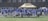 A group of football players wearing matching white and light blue uniforms stand in a line on a grassy field. They are holding helmets in their hands, and appear to be next to a beige tent structure. The background shows a wire fence and buildings.