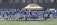 A group of football players wearing matching white and light blue uniforms stand in a line on a grassy field. They are holding helmets in their hands, and appear to be next to a beige tent structure. The background shows a wire fence and buildings.