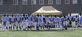 A group of football players wearing matching white and light blue uniforms stand in a line on a grassy field. They are holding helmets in their hands, and appear to be next to a beige tent structure. The background shows a wire fence and buildings.