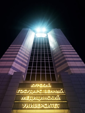 A tall, modern building illuminated at night with bright lights at the top. The structure has a symmetrical design with a grid pattern of windows running vertically. Neon yellow signs with Cyrillic text are displayed on the facade near the entrance, creating a striking contrast against the dark sky.