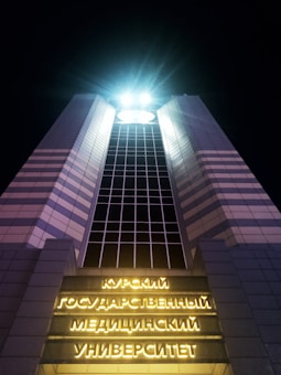 A tall, modern building illuminated at night with bright lights at the top. The structure has a symmetrical design with a grid pattern of windows running vertically. Neon yellow signs with Cyrillic text are displayed on the facade near the entrance, creating a striking contrast against the dark sky.