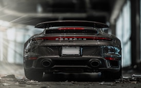 A sleek, high-performance sports car with a spoiler is parked in an indoor setting. The car's rear lights are illuminated, and the environment has an industrial vibe with concrete floors and large windows.