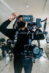 A person is operating a professional camera rig indoors. The individual is wearing dark clothing and a face mask, holding up a peace sign with one hand while adjusting the camera with the other. The background is slightly blurred with some office furniture visible.