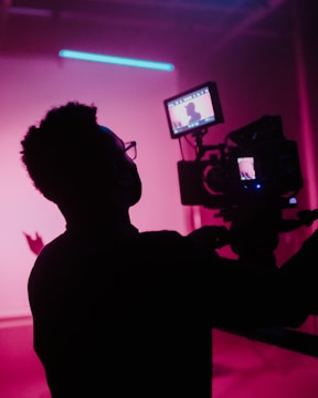 A silhouetted person operating a camera with a bright pink background, creating a dramatic and moody atmosphere. The focus is on the camera screen, which shows a blurred image of the scene being filmed. The scene is bathed in neon pink and purple lighting, adding a futuristic vibe.