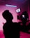 A silhouetted person operating a camera with a bright pink background, creating a dramatic and moody atmosphere. The focus is on the camera screen, which shows a blurred image of the scene being filmed. The scene is bathed in neon pink and purple lighting, adding a futuristic vibe.