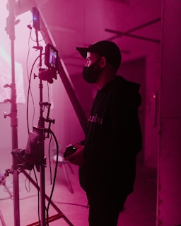 A person wearing a face mask and cap stands beside a film set, holding a device, possibly a camera. The scene is lit with soft pink lighting, casting a warm glow over the industrial setting. Various cables and filming equipment are visible in the background.