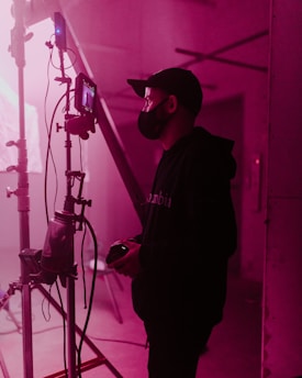 A person wearing a face mask and cap stands beside a film set, holding a device, possibly a camera. The scene is lit with soft pink lighting, casting a warm glow over the industrial setting. Various cables and filming equipment are visible in the background.