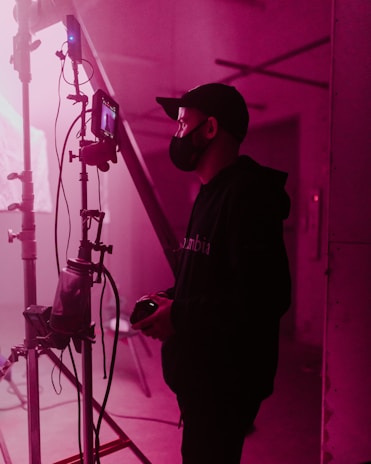 A person wearing a face mask and cap stands beside a film set, holding a device, possibly a camera. The scene is lit with soft pink lighting, casting a warm glow over the industrial setting. Various cables and filming equipment are visible in the background.