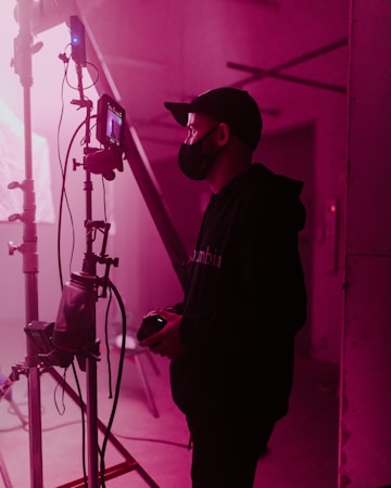 A person wearing a face mask and cap stands beside a film set, holding a device, possibly a camera. The scene is lit with soft pink lighting, casting a warm glow over the industrial setting. Various cables and filming equipment are visible in the background.
